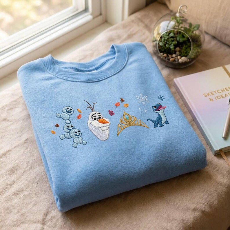 May include: A light blue crewneck sweatshirt featuring embroidered designs of a snowman, a stack of three small snow creatures, a gold crown, a blue lizard, and several small autumn leaves and snowflakes scattered across the chest area.