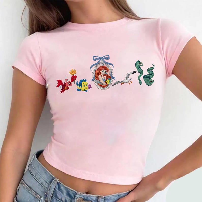 May include: A light pink short-sleeved t-shirt featuring a graphic print of characters from The Little Mermaid, including Sebastian, Flounder, Ariel in a framed oval portrait with a blue bow, Scuttle, and a green eel arranged in a horizontal row.