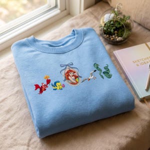 May include: A light blue crewneck sweatshirt featuring embroidered characters from The Little Mermaid, including a red crab, a yellow and blue fish, a central portrait of a mermaid in a frame, a white seagull, and a green eel.