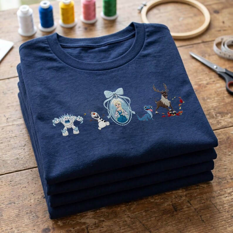 May include: A navy blue t-shirt featuring embroidered characters from the movie Frozen, including a white snow monster, a snowman, a portrait of a blonde woman in a blue dress with a bow, a blue lizard, and a brown reindeer with red leaves.