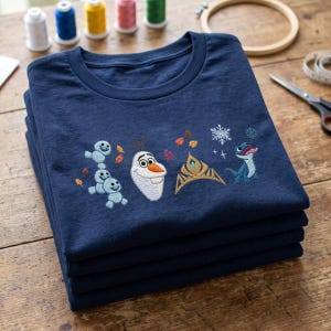 May include: A navy blue t-shirt featuring embroidered designs of a snowman, a group of small snow creatures, a golden crown, a blue lizard, and various colorful autumn leaves and white snowflakes scattered across the chest area of the garment.