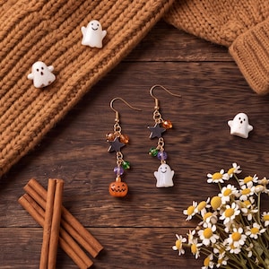 Könnte beinhalten: Ein Paar Ohrhänger mit goldfarbenen Haken, kleinen dunklen Sternen, bunten Perlen und Anhängern in Form eines weißen Geistes und eines orangefarbenen Kürbisses. Die Ohrringe sind zusammen mit losen weißen Geisterfiguren abgebildet.