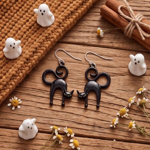 May include: A pair of black cat-shaped dangle earrings with silver-toned hooks. Each earring features a stylized feline silhouette with a curled tail and small green eyes. The earrings are displayed alongside small white ghost figurines.