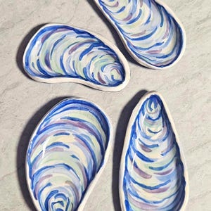 May include: Four ceramic oyster shell-shaped dishes. Each dish is painted with blue, purple, and green watercolour swirls, with a white border. The dishes are arranged on a light grey surface.