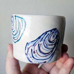 May include: A white ceramic cup featuring hand-painted blue and purple oyster shell motifs. The design consists of swirling, organic lines that mimic the texture of shells, distributed across the smooth, cylindrical surface of the vessel.