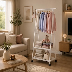 Puede incluir: Un perchero blanco en forma de A con ruedas que incluye una barra superior con varias camisas de manga larga en tonos pastel, un bolso pequeño y dos estantes inferiores con textiles doblados, un peluche y un bolso de mano beige.