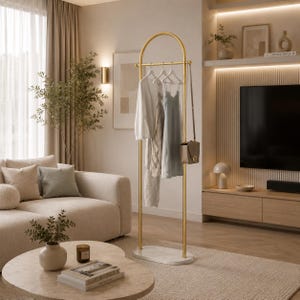 Puede incluir: Un perchero de metal dorado con una parte superior arqueada y una base ovalada blanca. El perchero cuenta con una barra horizontal para colgar ropa, que sostiene una blusa blanca de manga larga, una prenda de punto, un vestido y un bolso.