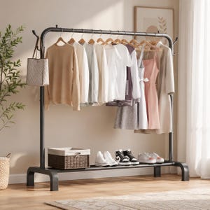 May include: A black metal garment rack with a top horizontal bar holding various shirts, dresses, and a tote bag on hangers. The base features a shelf containing a woven storage basket and four pairs of trainers arranged in a row.
