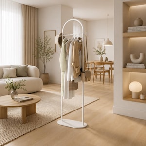 Puede incluir: Un perchero de metal blanco con un marco superior arqueado y una base ovalada. El perchero cuenta con una barra horizontal para colgar ropa, que actualmente sostiene varias prendas en tonos neutros, un sombrero y un pequeño bolso de hombro.