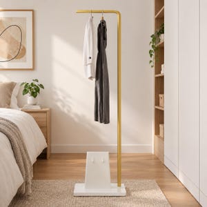 Op de afbeelding: Een goudkleurig metalen kledingrek met een horizontale hangstang en een witte rechthoekige voet. De voet is voorzien van een witte, trapeziumvormige steun met twee kleine haakjes. Een wit overhemd en een donkergrijze jurk hangen aan de stang.