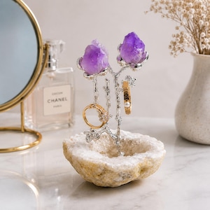 May include: A jewellery stand featuring a rough, circular white geode base with a silver-toned metal branch structure extending upward. Two raw purple amethyst crystals are attached to the top branches, holding a gold ring and a gold band.