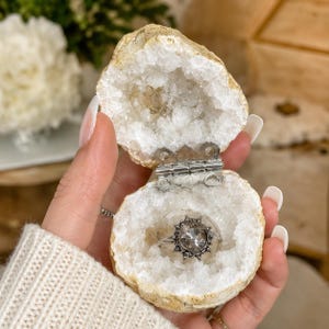 May include: A geode ring box featuring a natural, rough-textured exterior and a hollow interior lined with sparkling white quartz crystals. The two halves are connected by a silver-toned metal hinge, containing a silver ring with a round faceted stone.