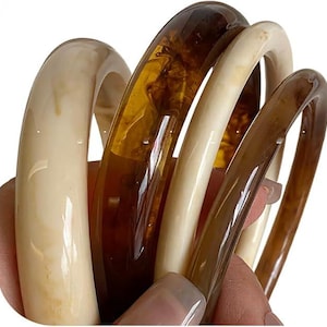 May include: A set of four smooth, rounded bangle bracelets in varying shades of cream, beige, and translucent amber. The bracelets feature a polished finish with marbled patterns, showcasing a mix of solid light tones and deep brown resin textures.