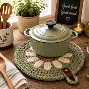 May include: A round, crocheted cotton trivet featuring a green background with a white daisy flower pattern in the center. The trivet includes a small brown leather tab with a metal rivet and a loop for hanging, positioned under a sage green pot.