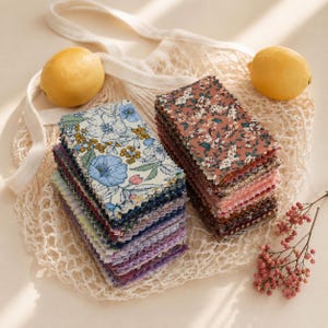 Può includere: Due pile di campioni di tessuto rettangolari con motivi floreali nei toni del blu, giallo e terracotta. I campioni sono disposti su una borsa a rete color crema insieme a due limoni gialli interi e un piccolo rametto di bacche rosa.