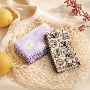 Puede incluir: Dos pilas de muestras de tela rectangulares que descansan dentro de una bolsa de malla de color crema. Una pila presenta un fondo morado claro con patrones de margaritas blancas, mientras que la otra muestra un fondo crema con ilustraciones de gatos y el texto LOVELY.