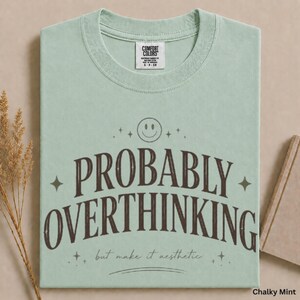 May include: A light mint green short-sleeve t-shirt with a crew neckline. The shirt features dark brown text that reads "PROBABLY OVERTHINKING" in a large arched font, with "but make it aesthetic" below it, a smiley face, and small stars.