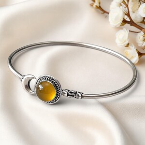May include: A silver bangle bracelet featuring a central round amber-coloured stone set in a twisted metal border. The design includes a crescent moon shape on one side of the stone and decorative metal wire wrapping on the opposite side of the band.
