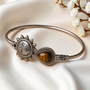 May include: A silver-toned metal bangle bracelet featuring a sun-shaped charm with etched details and a teardrop-shaped tiger's eye gemstone set in a twisted metal border. The bracelet has a smooth, polished finish and a simple open-ended design.