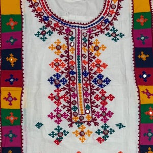 May include: A white tunic featuring vertical panels of multicolored fabric squares along the sides and a horizontal band at the hem. The garment is decorated with intricate, colorful geometric embroidery and small circular mirrors throughout.
