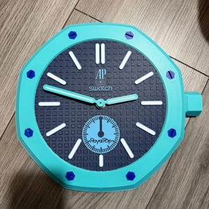 Puede incluir: Un reloj de pared turquesa con marco octogonal y detalles de tornillos azul oscuro. La esfera azul oscuro tiene un patrón de cuadrícula, marcadores de hora blancos y manecillas blancas. El texto AP, swatch y Royal Pop aparece sobre la subesfera.