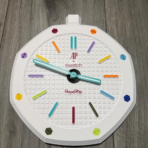 Puede incluir: Un reloj de pared octogonal blanco con una esfera de rejilla texturizada que incluye el texto AP x swatch y Royal Pop. El reloj tiene manecillas azul claro y marcadores de hora coloridos en tonos morado, naranja, amarillo, verde y azul.