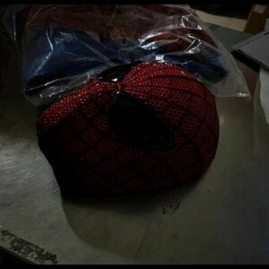 Puede incluir: Una m&aacute;scara roja con un patr&oacute;n de red negro y grandes lentes oculares angulares oscuros. La superficie de la m&aacute;scara est&aacute; cubierta con un dise&ntilde;o texturizado en forma de rejilla. Est&aacute; parcialmente envuelta en una bolsa de pl&aacute;stico transparente.