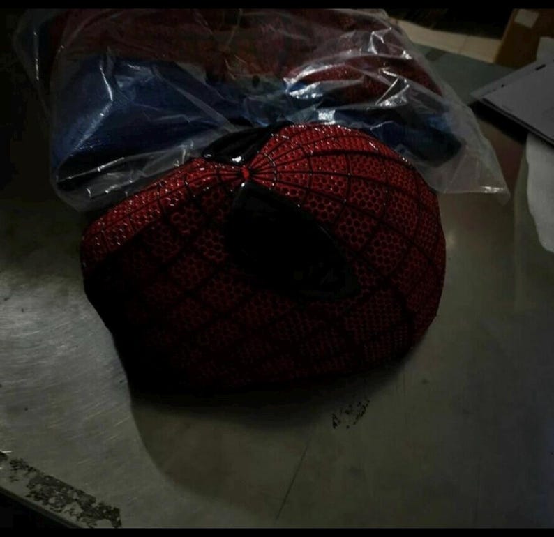 Puede incluir: Una m&aacute;scara roja con un patr&oacute;n de red negro y grandes lentes oculares angulares oscuros. La superficie de la m&aacute;scara est&aacute; cubierta con un dise&ntilde;o texturizado en forma de rejilla. Est&aacute; parcialmente envuelta en una bolsa de pl&aacute;stico transparente.