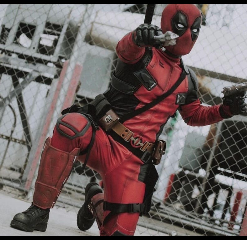 May include: A red and black tactical bodysuit featuring a full-face mask with white eye patches, black gloves, a utility belt with pouches, knee pads, and shin guards. The figure is in a kneeling pose, holding two handguns pointed forward.