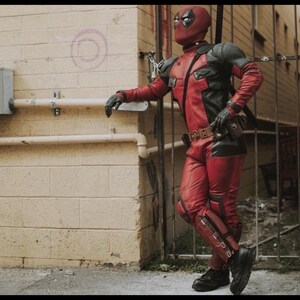 May include: A full-body costume featuring a red and black tactical suit with a matching mask, black gloves, and black boots. The suit includes shoulder armor, a utility belt with a central buckle, and leg guards, worn by a figure standing upright.
