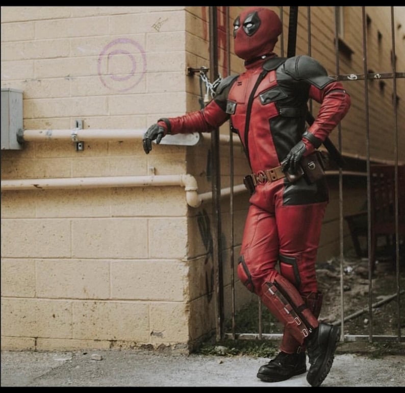 May include: A full-body costume featuring a red and black tactical suit with a matching mask, black gloves, and black boots. The suit includes shoulder armor, a utility belt with a central buckle, and leg guards, worn by a figure standing upright.