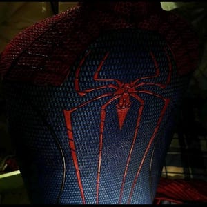 Puede incluir: Un traje de cuerpo completo texturizado en rojo y azul que presenta un prominente emblema de ara&ntilde;a roja centrado en la espalda. La tela tiene un patr&oacute;n de panal distintivo en toda la superficie, con secciones rojas en los hombros y un torso azul.