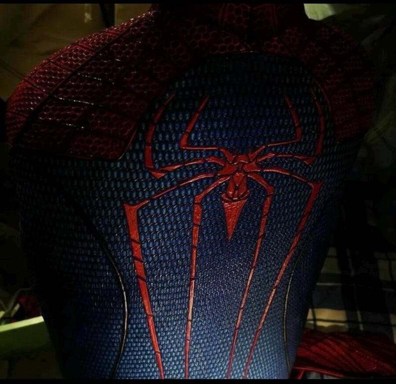 Puede incluir: Un traje de cuerpo completo texturizado en rojo y azul que presenta un prominente emblema de ara&ntilde;a roja centrado en la espalda. La tela tiene un patr&oacute;n de panal distintivo en toda la superficie, con secciones rojas en los hombros y un torso azul.