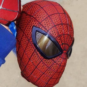 Puede incluir: Una m&aacute;scara roja de cabeza completa con un patr&oacute;n de telara&ntilde;a negro en relieve en la superficie. La m&aacute;scara incluye dos lentes oculares grandes y angulares con una textura de malla dorada met&aacute;lica oscura enmarcada por bordes negros gruesos.