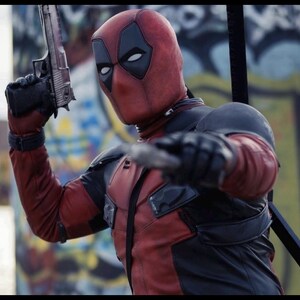 May include: A red and black textured full-head mask and tactical suit featuring black eye patches. The suit includes black shoulder pads and chest panels. One hand holds a metallic handgun pointed upward, while the other hand is extended forward.