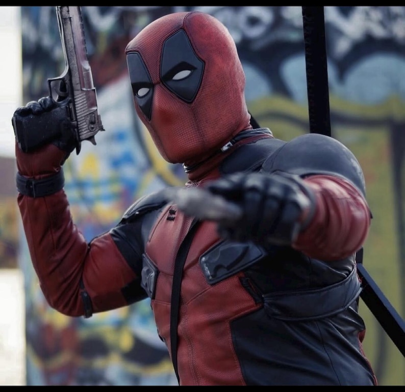 May include: A red and black textured full-head mask and tactical suit featuring black eye patches. The suit includes black shoulder pads and chest panels. One hand holds a metallic handgun pointed upward, while the other hand is extended forward.