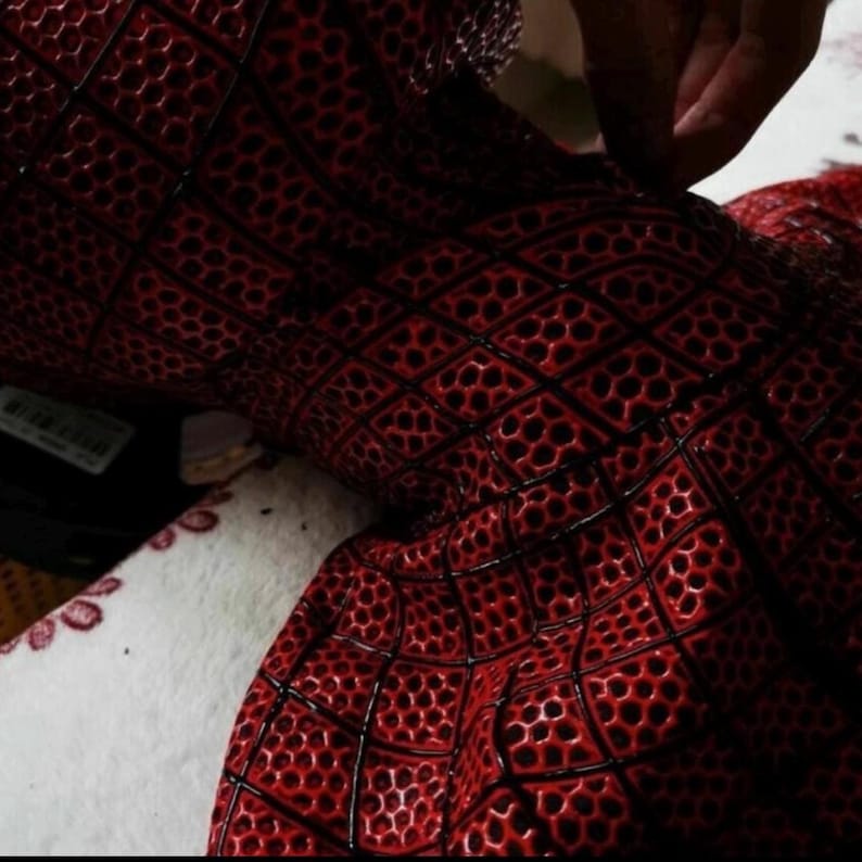 Puede incluir: Un tejido rojo con un patr&oacute;n de rejilla negro en relieve sobre una superficie texturizada similar a un panal. El material muestra un dise&ntilde;o geom&eacute;trico consistente con &aacute;reas rojas profundas y l&iacute;neas negras que forman cuadrados.