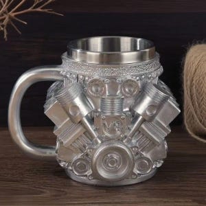 Könnte beinhalten: Ein silberfarbener Becher mit einem dreidimensionalen V8-Motorblock-Design. Die Außenseite zeigt geformte Kolben, Zylinder und mechanische Komponenten mit metallischem Finish. Der obere Rand besteht aus glattem Edelstahl mit einem stabilen Henkel.