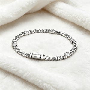 May include: A silver-toned chain link bracelet featuring a rectangular clasp engraved with the word PANDORA. The bracelet is accented with several clear, heart-shaped stones set at intervals along the polished metal chain links.