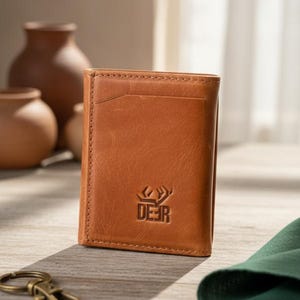 Puede incluir: Una cartera plegable de cuero color canela con una ranura vertical para tarjetas en la parte frontal y un logotipo grabado de una silueta de cabeza de ciervo sobre el texto DEER. La cartera tiene costuras visibles a lo largo de los bordes.