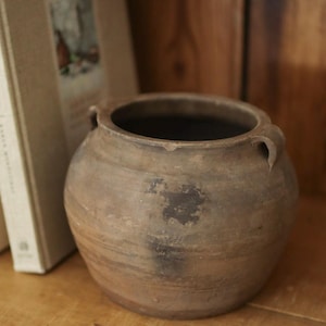 May include: A round, brown earthenware pot with a wide opening, a textured matte surface, and two small, curved handles on opposite sides. The vessel features subtle horizontal ridges and dark, weathered patches across its exterior body.