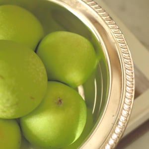 May include: A polished silver-toned metal bowl with a decorative rope-style rim, filled with several smooth, round, bright green apples. The bowl has a reflective metallic finish and a circular shape, containing the fresh fruit in a simple arrangement.