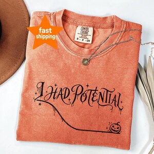 Peut inclure: Un t-shirt à manches courtes de couleur corail avec une encolure ronde. Le vêtement présente un texte graphique noir indiquant I HAD POTENTIAL, accompagné d'une petite toile d'araignée, d'une araignée et d'une citrouille d'Halloween.