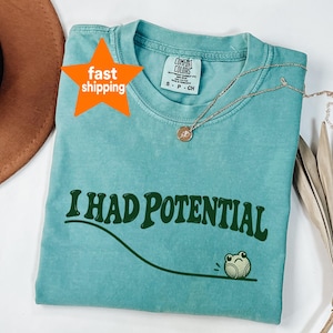 Op de afbeelding: Een blauwgroen T-shirt met korte mouwen en een ronde hals. Het shirt is voorzien van de donkergroene tekst "I HAD POTENTIAL" in een golvend lettertype boven een gebogen lijn en een kleine illustratie van een groen kikkertje dat er verdrietig uitziet.
