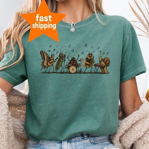 Może przedstawiać: Szałwiowo-zielony t-shirt z graficzną ilustracją zespołu owadów grających na instrumentach muzycznych, w tym motyla na gitarze, konika polnego śpiewającego, biedronki na perkusji, pszczoły na saksofonie i ślimaka na keyboardzie.