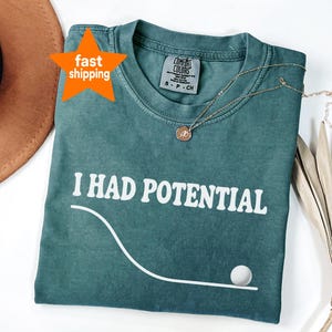 Peut inclure: Un t-shirt à col rond bleu sarcelle avec le texte blanc I HAD POTENTIAL imprimé au-dessus d'un graphique blanc représentant une ligne courbe et un petit cercle. Le t-shirt porte une étiquette de marque Comfort Colors et une étoile orange.