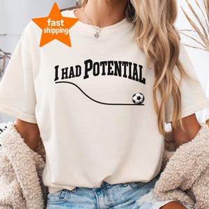 Puede incluir: Una camiseta de manga corta de color crema con el texto negro I HAD POTENTIAL en una fuente serif negrita. Debajo del texto hay una línea negra fina y curva que termina con un gráfico de un balón de fútbol en blanco y negro.