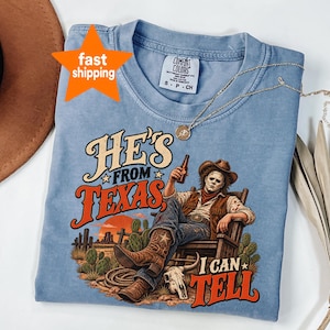 Pode incluir: Uma t-shirt de manga curta azul-clara com um gráfico de uma figura mascarada com chapéu de cowboy e colete sentada numa cadeira, segurando uma garrafa. O texto diz HE'S FROM TEXAS I CAN TELL. Etiqueta da marca Comfort Colors.