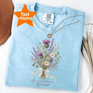 May include: A light blue short-sleeve t-shirt with a crew neckline and a Comfort Colors brand label. The centre features a printed bouquet of purple thistles, white daisies, and greenery tied with a beige ribbon, with the text be a good human.