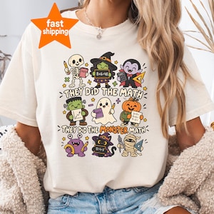 Op de afbeelding: Een crèmekleurig t-shirt met een afbeelding van diverse cartoonmonsters die bezig zijn met schoolactiviteiten. De tekst luidt "THEY DID THE MATH" en "THEY DID THE MONSTER MATH" naast illustraties van een skelet, heks, vampier, Frankenstein, geest, pompoen, paars monster, kat en mummie.